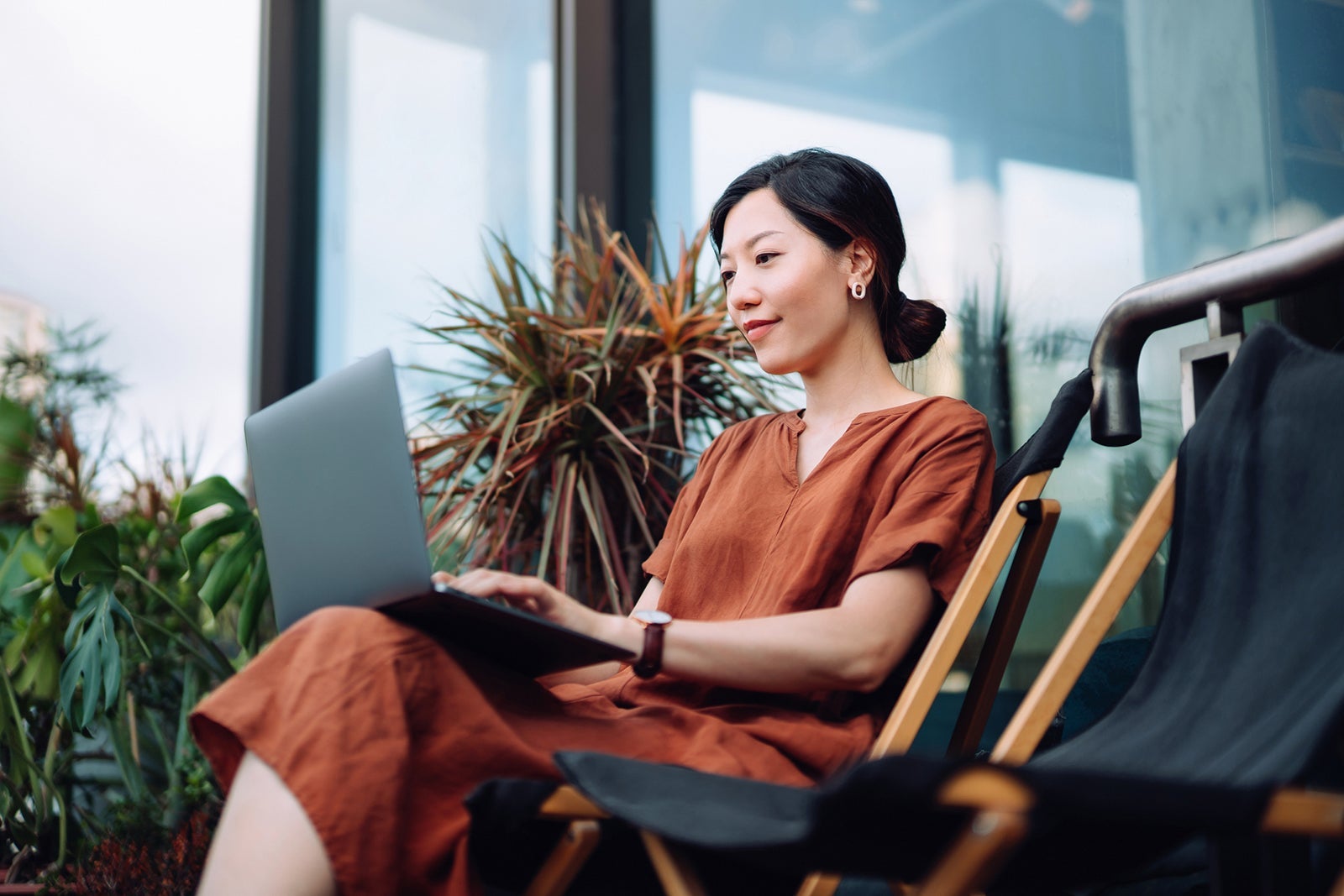 Young Asian woman sitting on deck chair in the balcony, working on laptop on lap at home. Surrounded by beautiful houseplants. Working from home, freelance concept. Lifestyle and technology