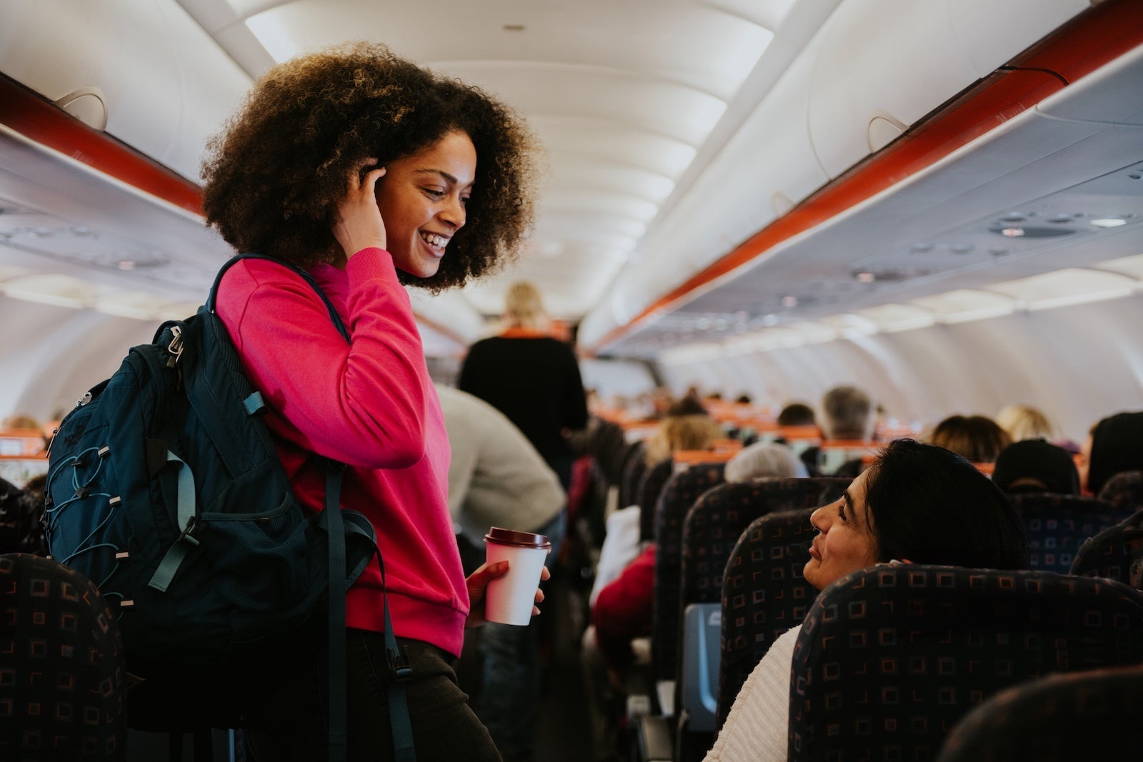 A woman with a backpack boards a small commercial aircraft, greeting a woman who is already seated on the plane